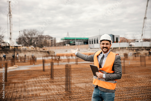 A cheerful engineer is standing on site in building process with tablet in his hands and showing at site while smiling at the camera.