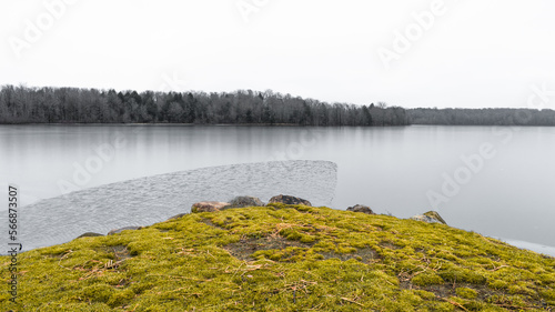 Lake reservoir on cold day with stony grass lawn in foreground and pine forest in the distance.