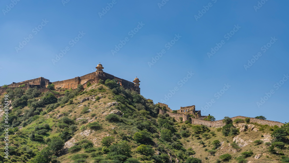 An ancient defensive fortress wall runs along the ridge of the mountain. Watchtowers with domes are visible against the blue sky. Green vegetation on the slopes. India. Amber Fort. Jaipur