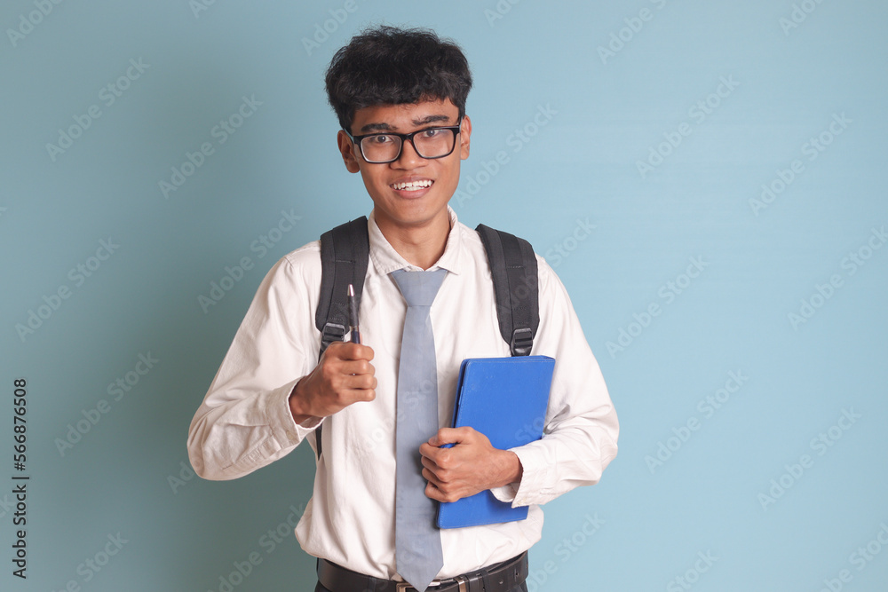Indonesian senior high school student wearing white shirt uniform with gray tie holding some books, smiling and looking at camera. Isolated image on blue background Indonesian senior high school student wearing white shirt uniform with gray tie holding some books, smiling and looking at camera. Isolated image on blue background