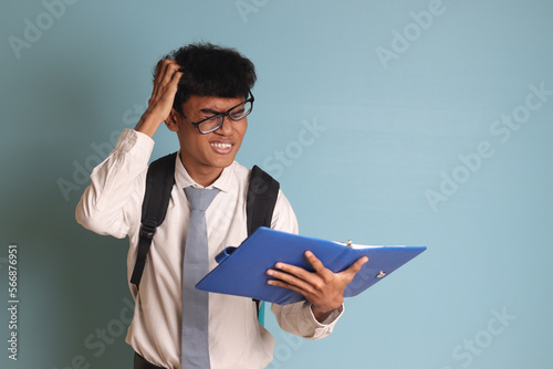 Indonesian senior high school student wearing white shirt uniform with gray tie writing on note book using pen and thinking about an idea. Isolated image on blue background photo