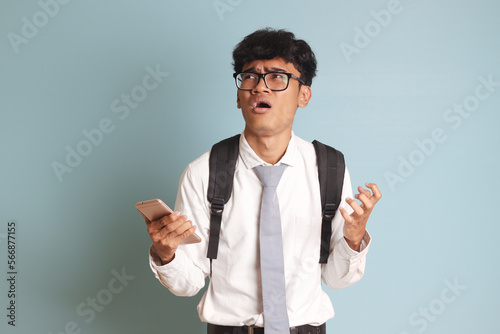 Indonesian senior high school student wearing white shirt uniform with gray tie feeling disappointed making angry hand gesture while holding mobile phone. Isolated image on blue background photo