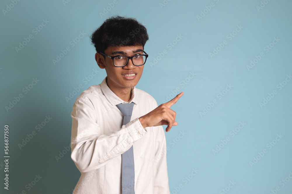Indonesian senior high school student wearing white shirt uniform with gray tie showing product, pointing at something and smiling. Isolated image on blue background Indonesian senior high school student wearing white shirt uniform with gray tie showing product, pointing at something and smiling. Isolated image on blue background