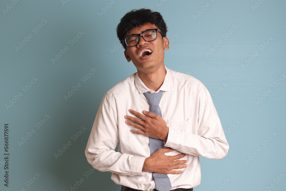 Indonesian senior high school student wearing white shirt uniform with gray tie laughing loudly while holding his chest. Fun comedy concept. Isolated image on blue background