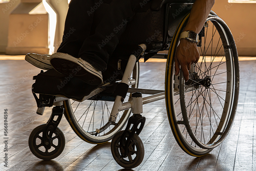 close em pernas e mão masculinas de um cadeirante em sua cadeira de rodas em um piso de tabua corrida e luz do sol ao fundo