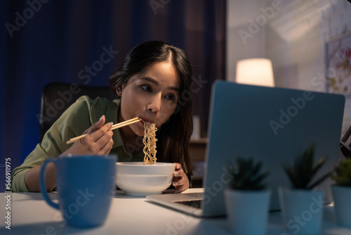Portrait of Asian businesswoman eating instant noodles while working on laptop in living room at home at night.