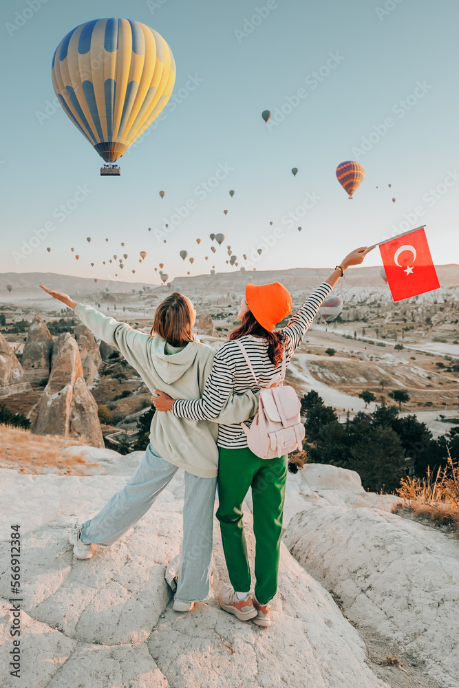 Girl friends travellers with turkish flag, hugging on a viewpoint and admiring view of flying hot air balloons in Cappadocia