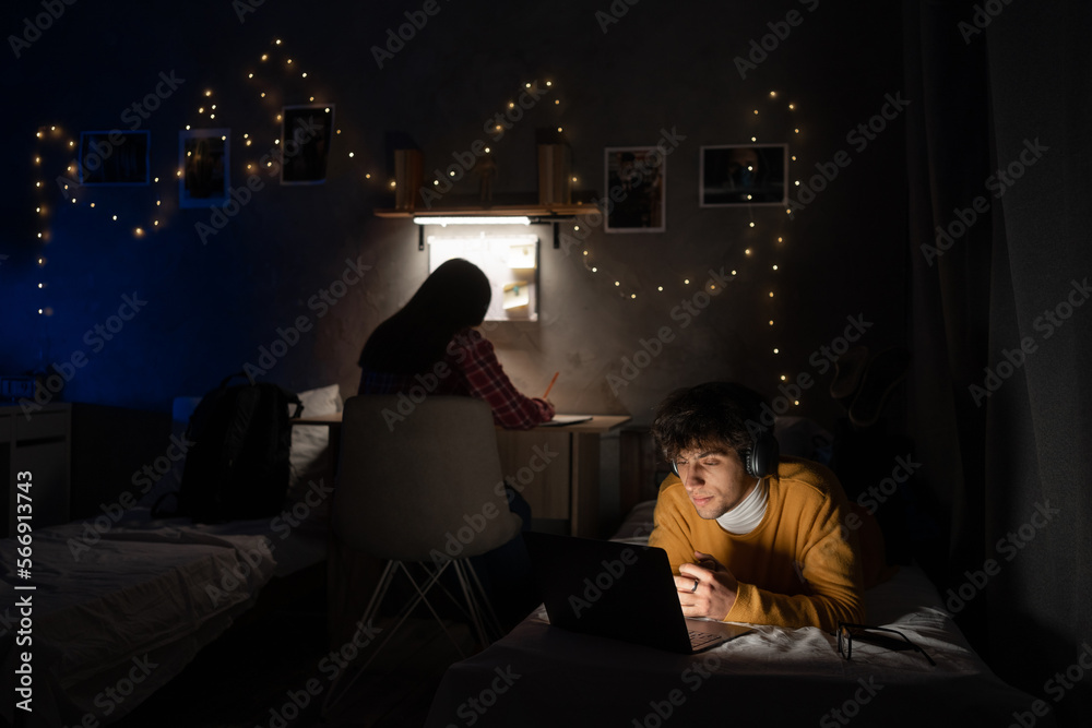 Male college student wearing headphones lies on bed in shared house working on laptop and girl while studying on background.