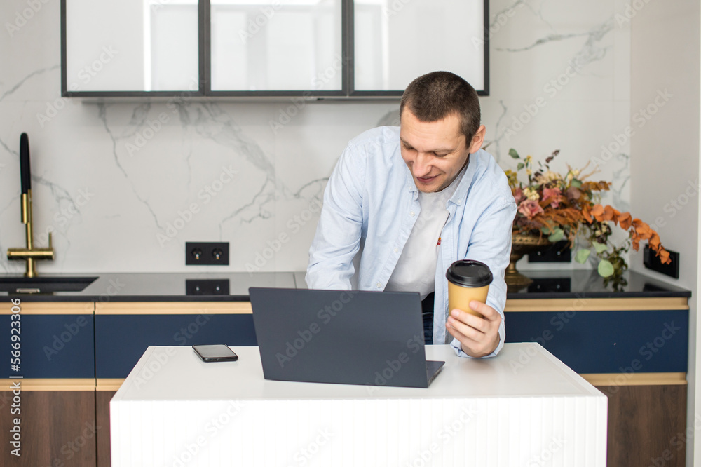 A young man standing in the kitchen is working on a laptop and holding a disposable cup A young man standing in the kitchen is working on a laptop and holding a disposable cup