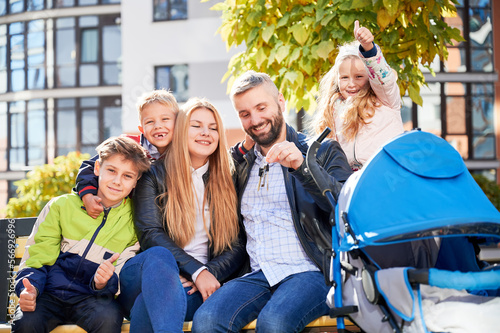 Happy family - father, mother and children having fun together on playground. Smiling father holding keys from new apartment, kids showing thumbs up. Modern residential buildings on background. © anatoliy_gleb