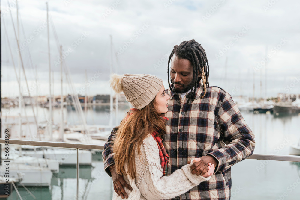 Fototapeta premium a multiethnic couple looking at each other holding hands, the harbor in the background