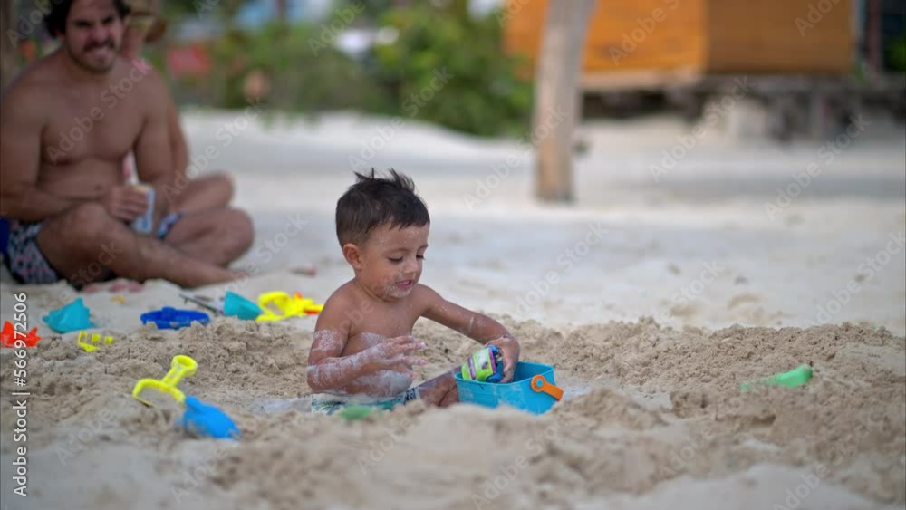 Slow motion of a little baby boy sitting in a hole on the beach playing with beach toys while a woman pours water into it