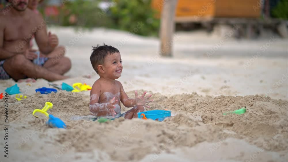 Slow motion of a cute little toddler exited and smiling while a woman pours water into a hole made on the beach to play Slow motion of a cute little toddler exited and smiling while a woman pours water into a hole made on the beach to play