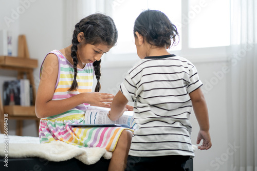 Young girl sister sitting with cute kid innocent brother enjoy reading book for him in bedroom at home. Portrait of adorable Asian girl who loves and cares for her little boy preschool sibling.