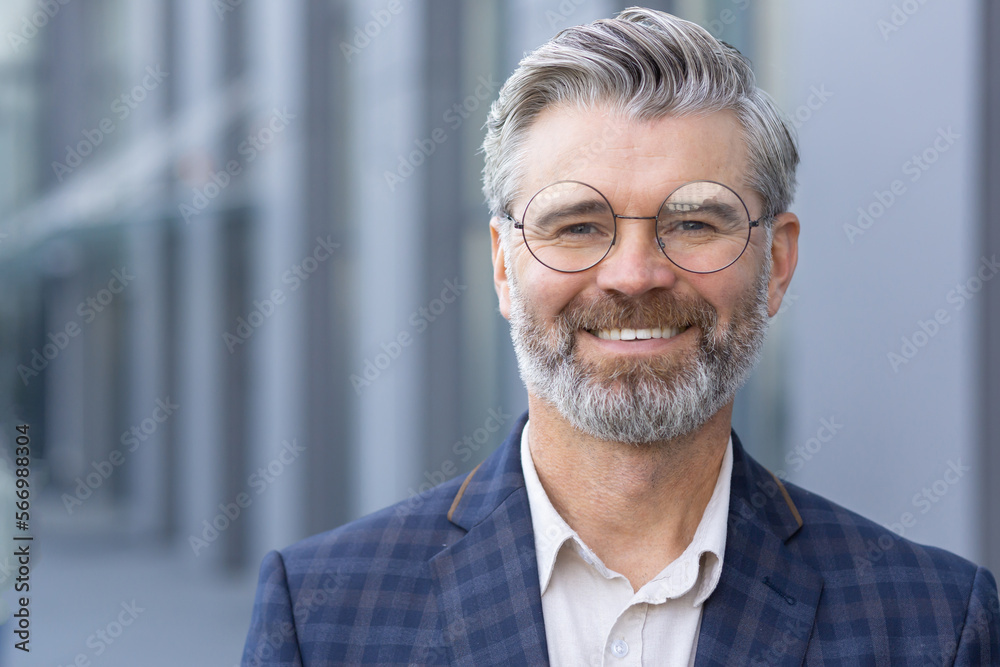 Successful gray haired man in business suit close up, portrait of mature businessman with beard and glasses, banker investor smiling and looking at camera from outside office building boss satisfied. Successful gray haired man in business suit close up, portrait of mature businessman with beard and glasses, banker investor smiling and looking at camera from outside office building boss satisfied.