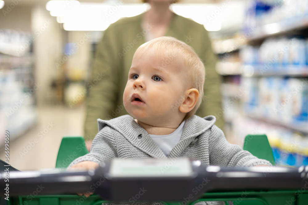 Mother pushing shopping cart with her infant baby boy child down department aisle in supermarket grocery store. Shopping with kids concept Mother pushing shopping cart with her infant baby boy child down department aisle in supermarket grocery store. Shopping with kids concept