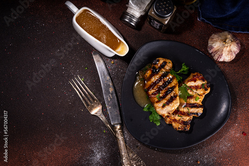 American bbq food concept. Grilled barbeque pork meat steaks with traditional gravy sauce, spices and greens, on plate board, dark background top view copy space 