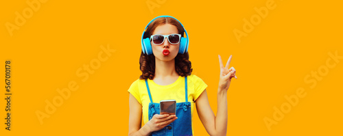 Portrait of stylish young woman in headphones listening to music with smartphone on yellow background