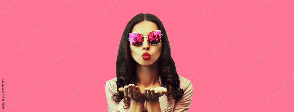 Portrait of beautiful young woman blowing her lips sending air kiss posing wearing sunglasses, leather jacket on pink background