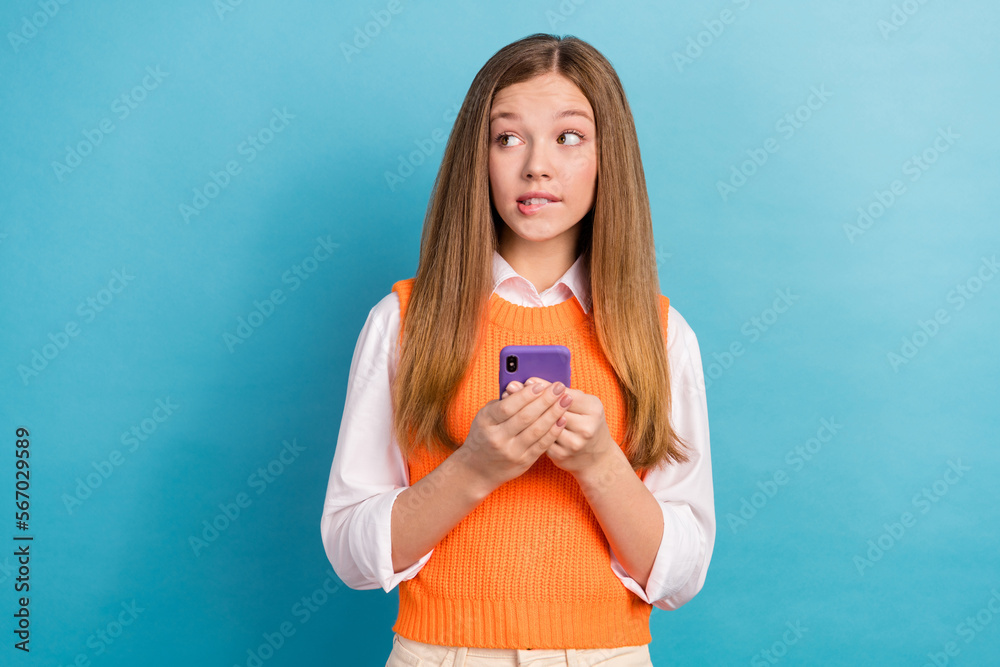 Photo of minded pensive girl biting lips hold telephone look empty space isolated on blue color background Photo of minded pensive girl biting lips hold telephone look empty space isolated on blue color background