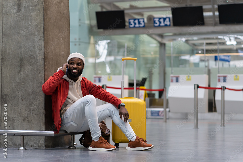 Φωτογραφία Cheerful black male sitting on floor relaxing near wall airport with before announcement for flight check-in, talking on smartphone, waiting friend for together journey