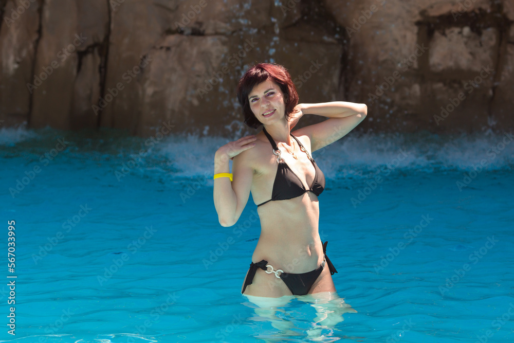 An adult brunette woman in a black swimsuit smiling stands in the water at the waterfall. An adult brunette woman in a black swimsuit smiling stands in the water at the waterfall.