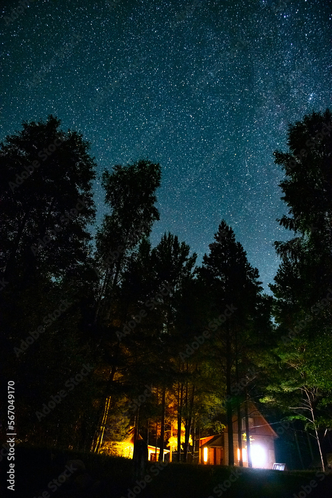 Picturesque starry sky above the earth.An atmospheric night in a hut,alone with nature.Fantastic panorama of the landscape with a wooden house in a pine forest against the background of the starry sky
