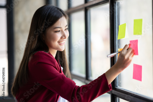 Smart asian business woman mentor coach leader writing idea or task on post it sticky notes on glass wall.