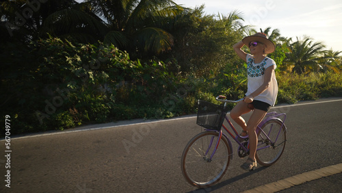 Pretty mature senior woman holding her hat while biking away from the sun, backlit, wearing sunglasses in a tropical setting.