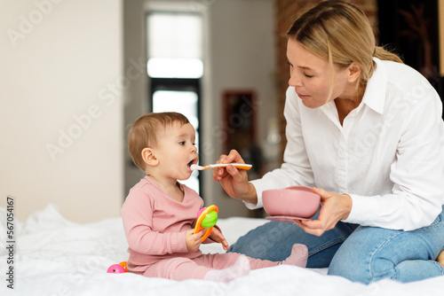 Childcare. Loving mother feeding her cute baby girl from spoon, sitting on bed, caring mom giving healthy porridge