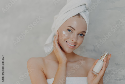Close up shot of pretty young European woman with minimal makeup  applies moisturising cream on face  smiles gently and looks at camera  wears bath towel on head  isolated over grey background