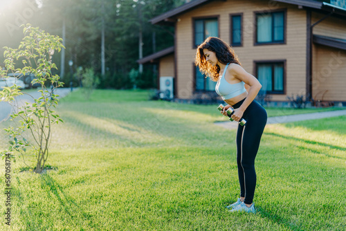 Full length shot of active sporty young woman raises dumbbells has workout outdoor near house stands on green lawn, dressed in sportsclothes, tries to be in good shape, stays fit and healthy