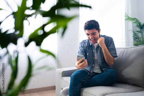 Hispanic teenager boy sitting on couch and celebrating good news on phone.