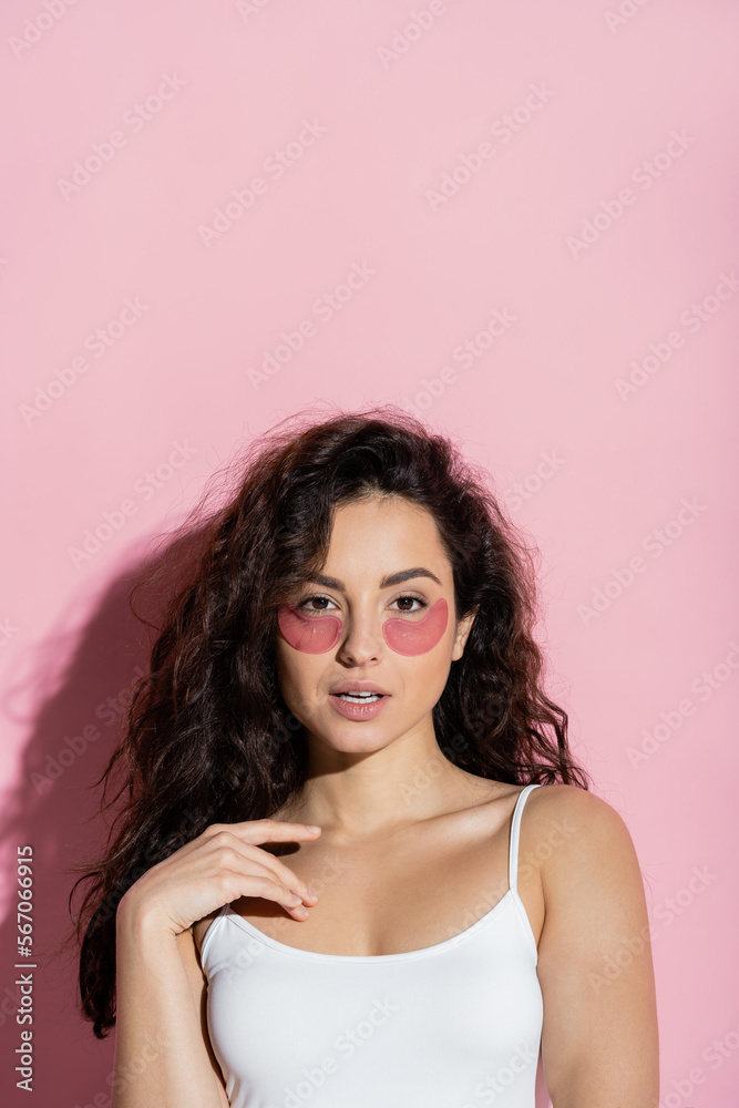 Curly brunette woman with hydrogel eye patches touching chest on pink background.