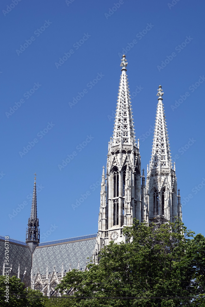 Two high spires on Votivkirche and church roof rise above green tree branches in the central part of Vienna