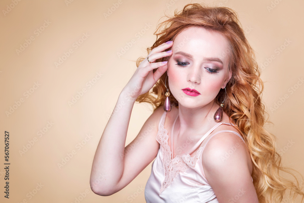 Beautiful red-haired girl with wavy hair, make-up, in a silk top and long earrings posing on a beige background in the studio. Woman touching her face and looking down Beautiful red-haired girl with wavy hair, make-up, in a silk top and long earrings posing on a beige background in the studio. Woman touching her face and looking down