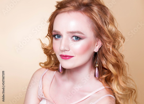 Beautiful red-haired girl with wavy hair, make-up, in a silk top and long earrings posing on a beige background in the studio. Woman looking at the camera and smiling