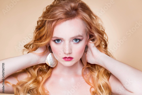 A beautiful red-haired girl with wavy hair and makeup poses on a beige background in the studio in round gold earrings. Woman holding her hands on the back of her head and looking at the camera
