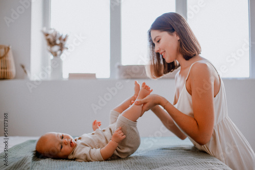 Caring young mother kissing baby's feet while making gymnastics on bed at home, side view, copy space. Loving mom spending time with her cute little daughtter