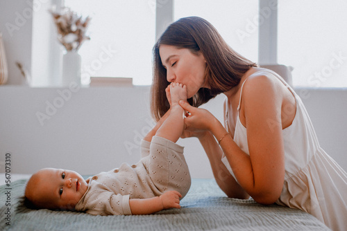 Caring young mother kissing baby's feet while making gymnastics on bed at home, side view, copy space. Loving mom spending time with her cute little daughtter