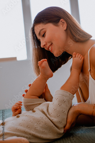 Caring young mother kissing baby's feet while making gymnastics on bed at home, side view, copy space. Loving mom spending time with her cute little daughtter
