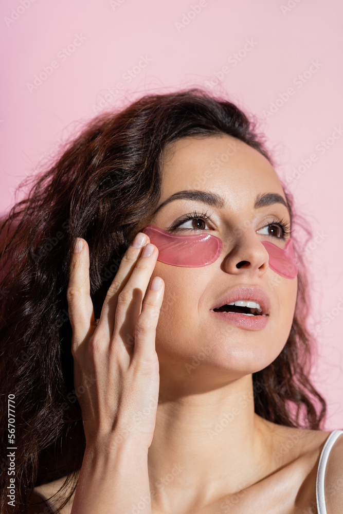 Fototapeta premium Young brunette woman applying hydrogel eye patch on face on pink background.