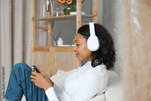Side view portrait of a happy african amercian girl listening music,podcast or lecture on sofa at home with phone in her hands. Online application and modern technologies concept.