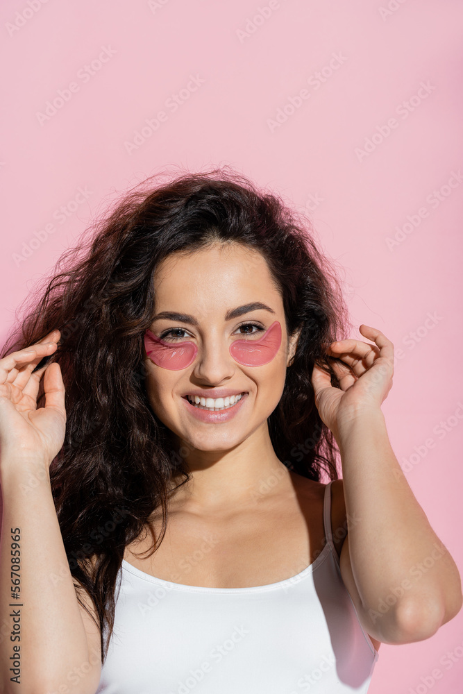 Fototapeta premium Cheerful young woman with hydrogel patches touching hair on pink background.