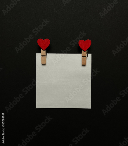 Empty white sticker, wooden clothespins with red hearts on black background, flat lay.