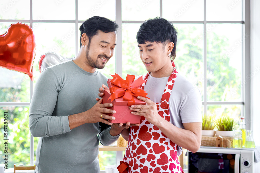 Happy smiling LGBT couple sharing sweet moment together on Valentine Day at kitchen Asian gay male giving red gift box present to lover, celebrating memorable anniversary unforgettable romantic dating