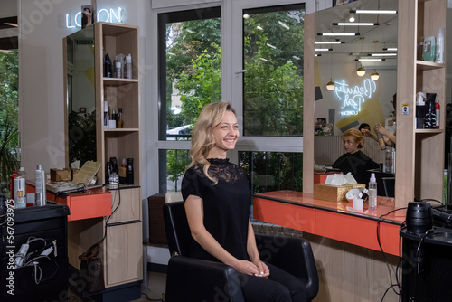 Blonde woman in black blouse sits on chair near workplace of makeup artist with large mirror. Person smiles looking contentedly in modern beauty salon © SlavaStock