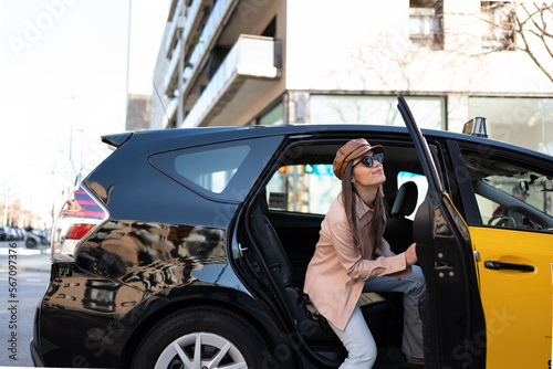 girl getting out of a cab, smiling woman with sunglasses, leather beret, pink blazer and jeans, modern and very attractive caucasian. In the background office buildings © Cristina