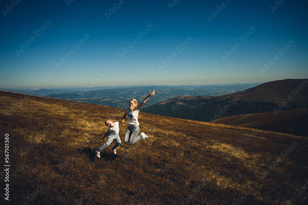 Obraz premium Happy family, mother and daughter, in summer clothes, having fun and laughing, holding hands and pulling each other on top of a mountain, on a sunny day in summer.