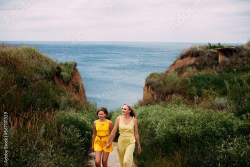 A beautiful family, a mother and daughter in yellow summer clothes walk hand in hand and smile, among the hills near the ocean on a bright sunny day. © Ihor